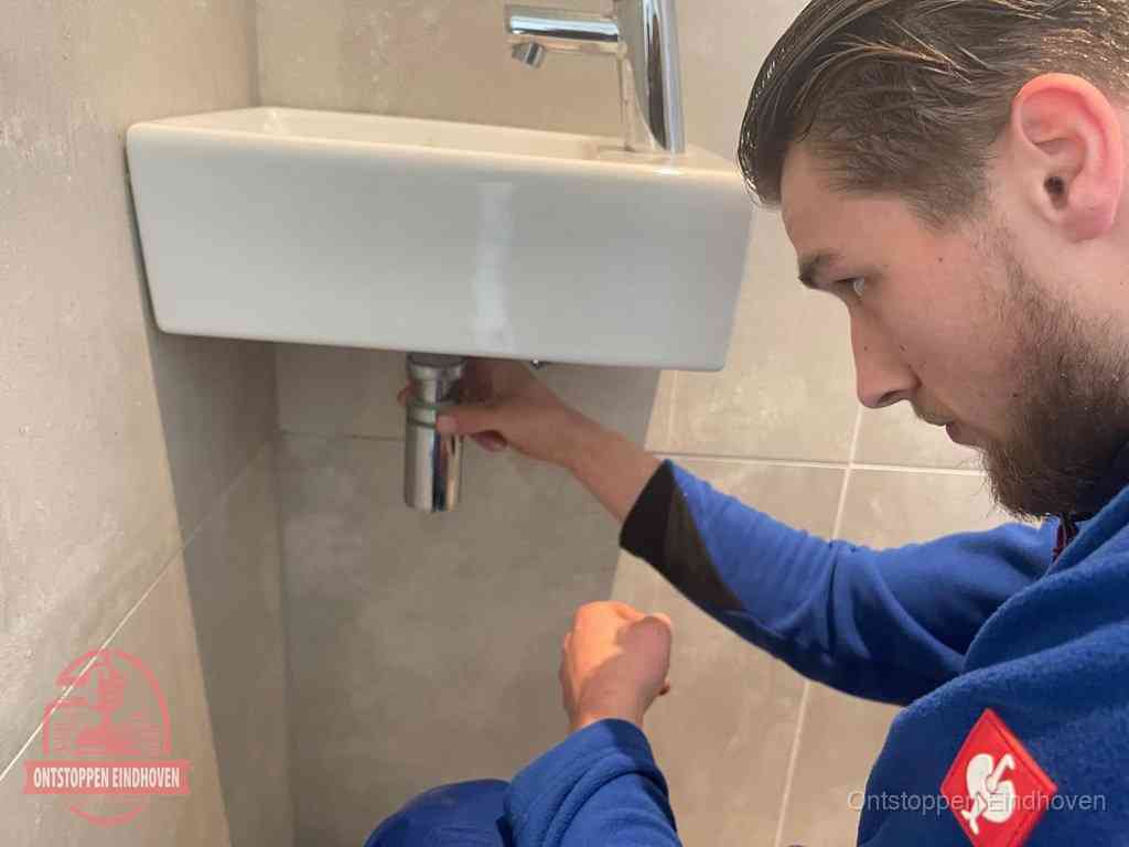 Een monteur draait met de hand de zwanenhalssifon onder een fonteinwasbak in de toiletruimte los om de bron van rioollucht in Eindhoven te vinden Een monteur draait met de hand de zwanenhalssifon onder een fonteinwasbak in de toiletruimte los om de bron van rioollucht in Eindhoven te vinden